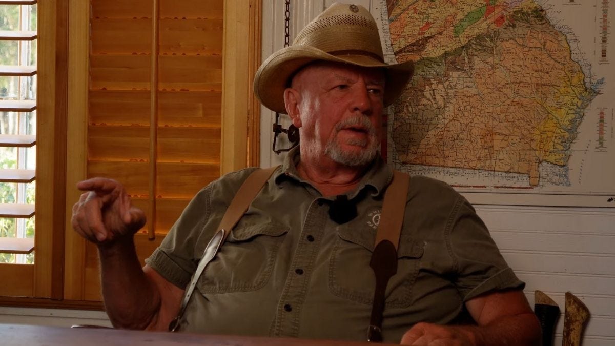 A farmer wearing a straw hat and suspenders speaks inside a wooden-paneled room at White Oak Pastures, with a vintage map of Georgia hanging on the wall behind him. A farmer wearing a straw hat and suspenders speaks inside a wooden-paneled room at White Oak Pastures, with a vintage map of Georgia hanging on the wall behind him.