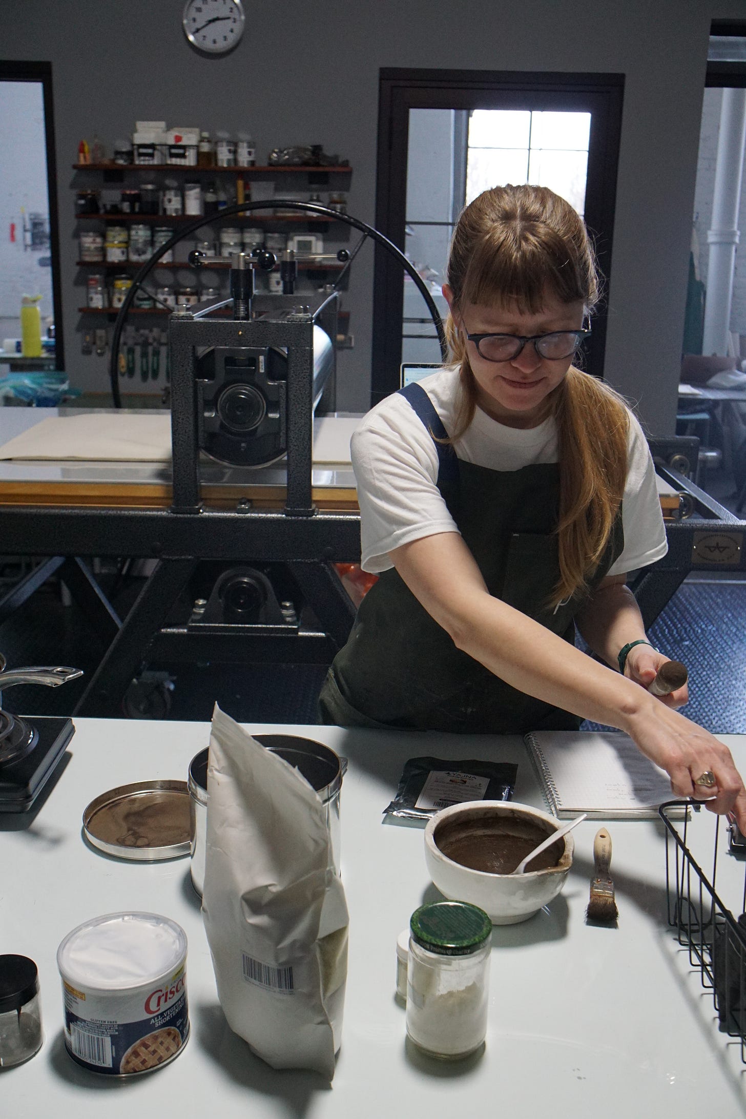 A white woman with long blonde hair and a green apron reaches out of frame. On the work surface in front of her, there rests a variety of bowls and containers.