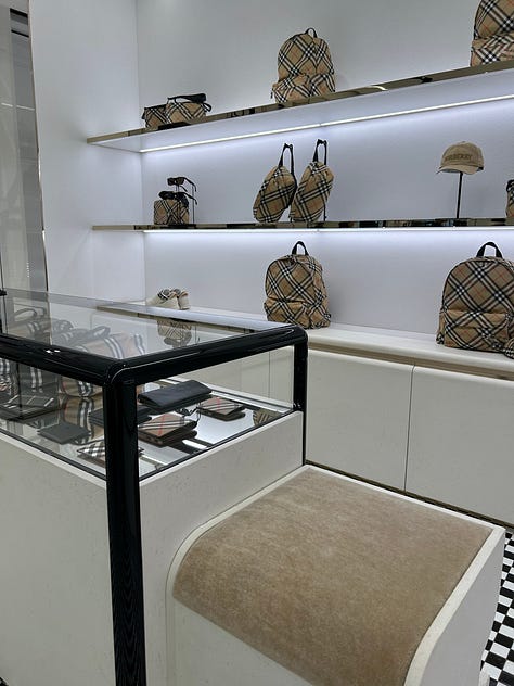 Three-photo gallery from an Intern Pierre primary channel-check store visit to a Burberry flagship, documenting the gap between the company's articulated heritage refocus narrative and the actual visual merchandising on the floor. Left photo: the women's handbag wall positioned at the storefront entry zone, with structured leather Burberry shoulder bags, hobo bags, and check-pattern canvas backpacks displayed on backlit white wall shelves above a glass jewelry display case and a beige suede storage bench. Center photo: a sparse men's ready-to-wear central display featuring two mannequins in muted olive and grey tailored outfits styled with bucket hats, flanked by half-empty wood-tone clothing rails and a minimalist wood bench, occupying the deep interior of the store far from the entrance. Right photo: men's outerwear and ready-to-wear racks in a back-of-store section, featuring a Burberry check-pattern bomber jacket with contrast sleeves, a red and beige check overshirt, and additional checked outerwear hanging on chrome rails with folded scarves and a leather portfolio bag arranged on a low display plinth. The sequence demonstrates the structural mismatch between the company's "hero categories take prominence" merchandising claim and the actual layout, where handbags and accessories anchor the entry zone, men's outerwear and trench coats require navigating deep into the store, and check-pattern saturation remains heavy across ready-to-wear and outerwear categories despite the public narrative of a quiet-luxury heritage repositioning. Channel-check evidence relevant to Burberry $BRBY.L investors evaluating the credibility of the current turnaround thesis and the alleged return to outerwear and scarves as the brand's anchor categories under CEO Joshua Schulman.