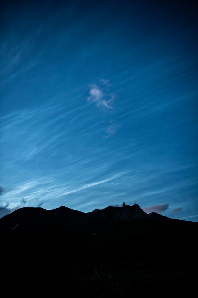 A deep blue night sky streaked with pale, rippled noctilucent cloud rises above a black silhouetted ridge, a jagged rock spire breaking its skyline. A small tuft of cloud near the center catches faint pink light.