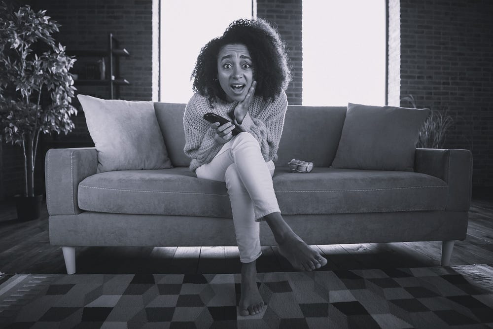Black woman on a sofa with phone in one hand, the other hand is pressed against her face in an expression of horror and surprise. Black woman on a sofa with phone in one hand, the other hand is pressed against her face in an expression of horror and surprise.