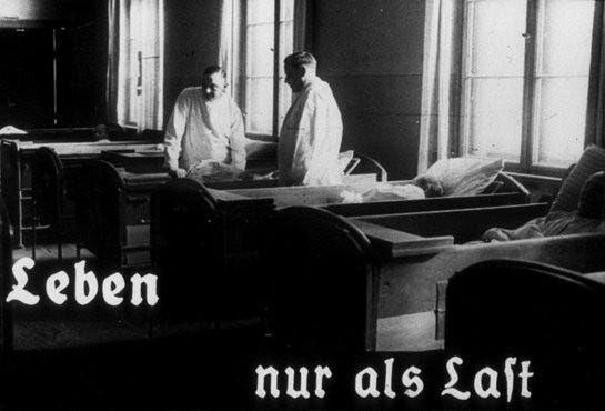 Two men stand among several occupied wooden beds wearing white medical uniforms. White text along the bottom read, “Leben nur als Last” which translates to, “Life only as a burden.”