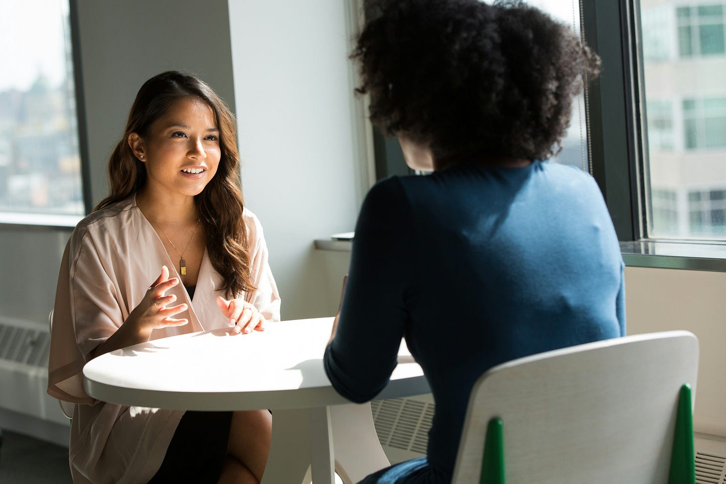 Two women sitting at a table, speaking. One is facing the camera, the other shows her back. Photo by Christina(at)wocintechchat(dot)com via Unsplash.