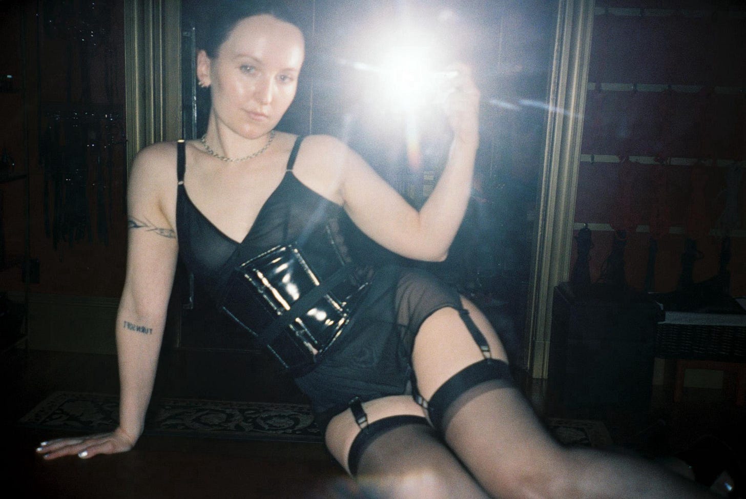 A film photo of Evyn Silver, a white femme with dark hair worn up in a twist, taking a selfie in a mirror. She's sitting on a leather table leaning on one arm with her legs bent to the side. She's wearing a black mesh set with suspenders, stockings, and a shiny black corset. The flash from the camera obscures most of the background except for a few rows of rope and equipment hanging on the walls.