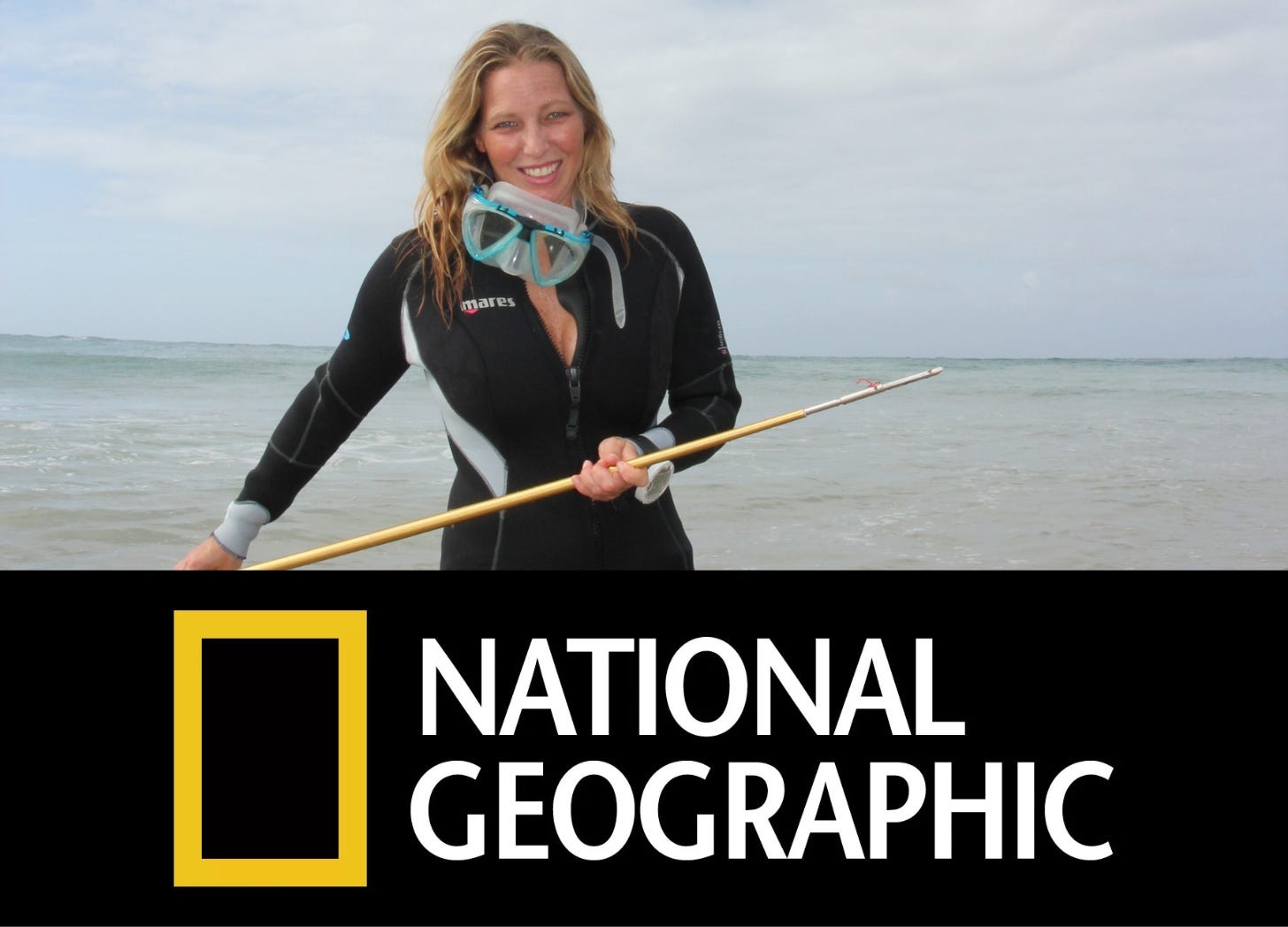 A white woman with long blond hair is wearing a black scuba suit and holding a spear. She is standing in the ocean with a blue sky behind. 