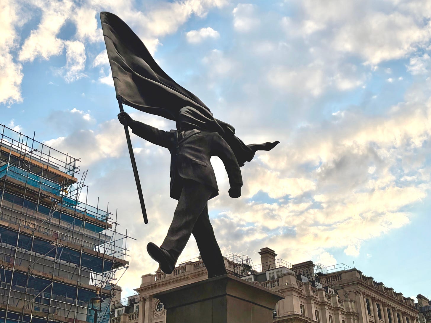 Banksy's statue in London of a suited man striding off a plinth, carrying a flag on a pole whose fabric streams back and wraps across his head, hiding his face. Banksy's statue in London of a suited man striding off a plinth, carrying a flag on a pole whose fabric streams back and wraps across his head, hiding his face.