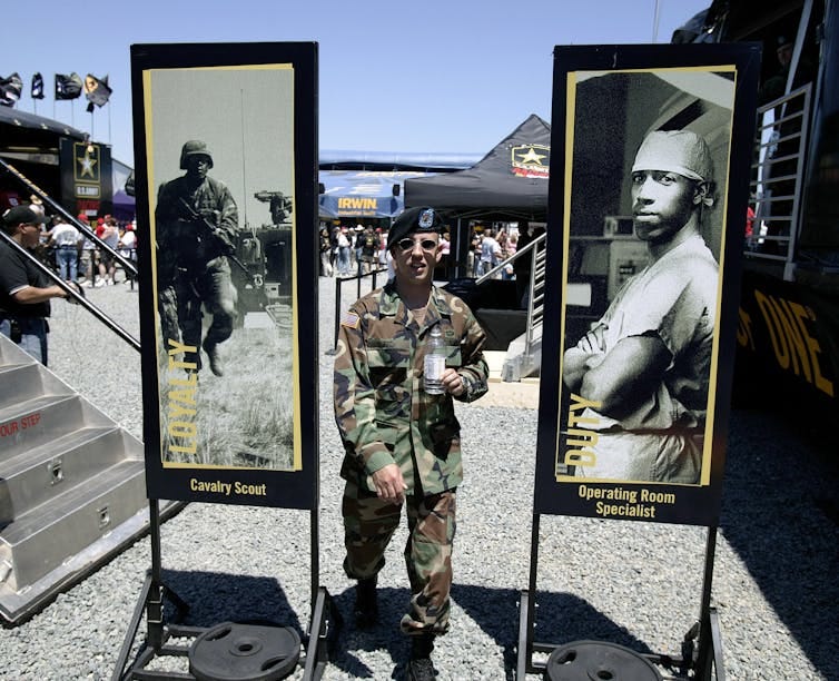 An Army recruiter dressed in military garb stands between two posters depicting Black men in the armed forces.