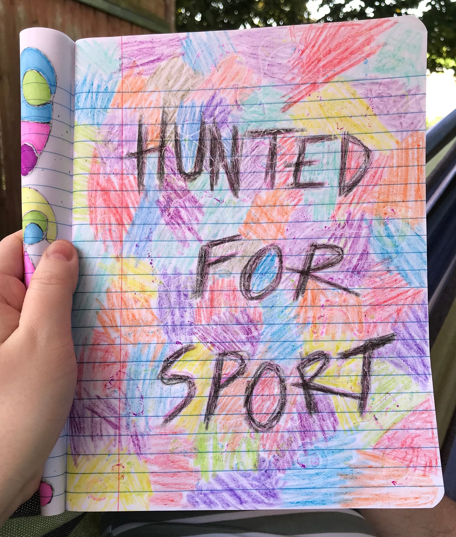 A sketch made with crayons on notebook paper that says “HUNTED FOR SPORT” in messy black writing. The background is filled in with multicoloured scribbled patches.