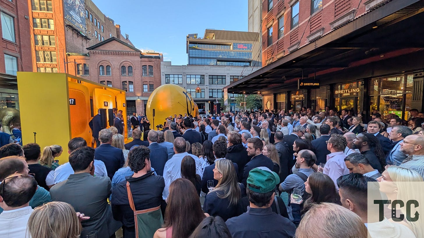 Former NFL quarterback Eli Manning addresses a large crowd at the Breitling NFL launch event. Behind him, an oversized yellow backdrop featuring numbered lockers and a giant inflatable football dominates the outdoor setting.
