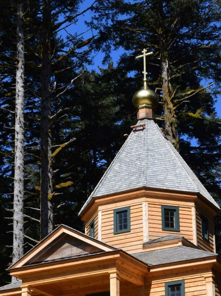 Golden onion dome topped with a cross on an octagonal wood-shingled cupola, seen through evergreen trees against a blue sky.