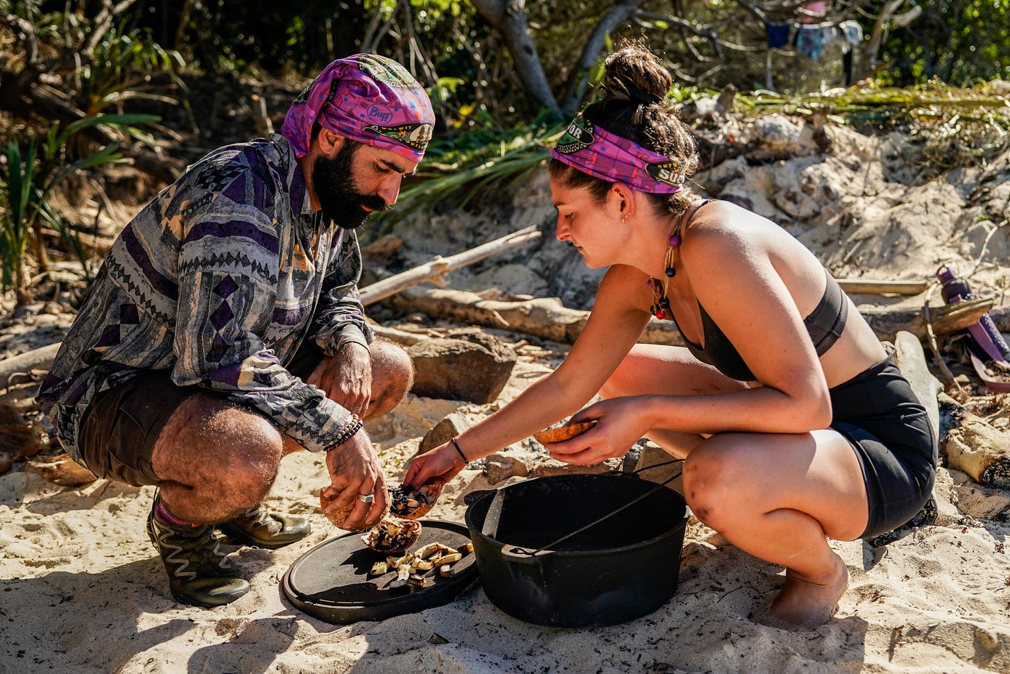 Bianca and a castmate crouch in the sand, seeming to prep some food.