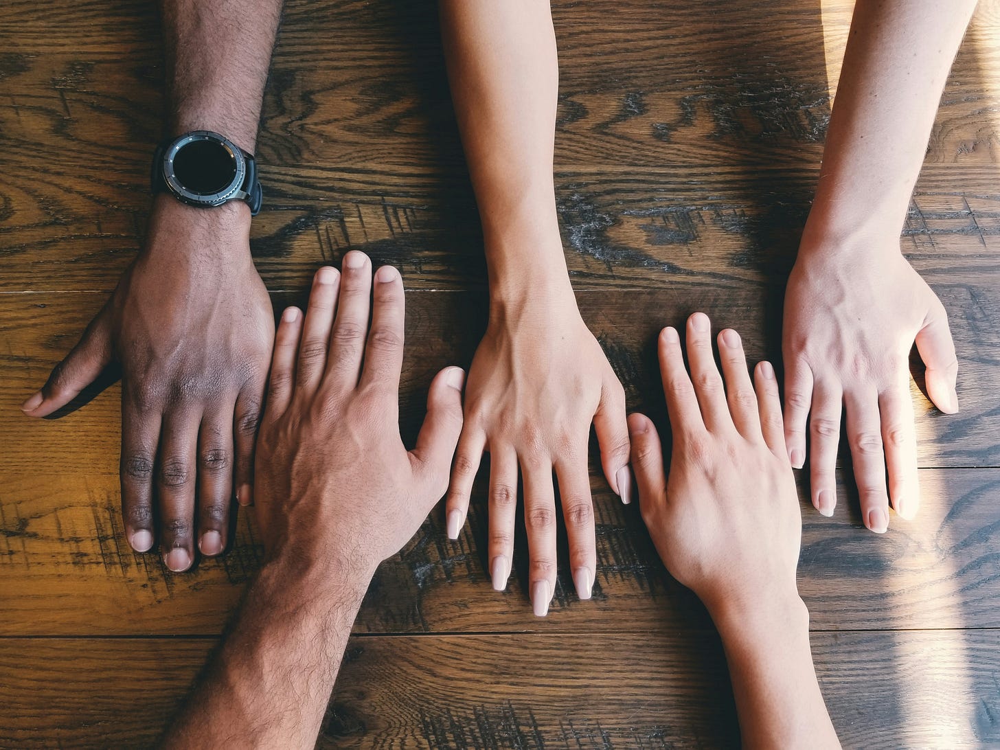 Looking down at a wooden table where five hands are reaching in from top and bottom—different colors, different genders—to meet in one equal row across the table. Looking down at a wooden table where five hands are reaching in from top and bottom—different colors, different genders—to meet in one equal row across the table.