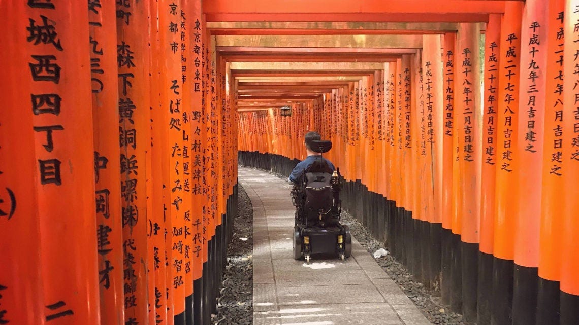 A power wheelchair user travels through the iconic orange torii gate tunnel at Fushimi Inari Shrine in Kyoto, Japan, with hundreds of vermilion gates lined with black bases and Japanese text stretching into the distance along a paved pathway. A power wheelchair user travels through the iconic orange torii gate tunnel at Fushimi Inari Shrine in Kyoto, Japan, with hundreds of vermilion gates lined with black bases and Japanese text stretching into the distance along a paved pathway.