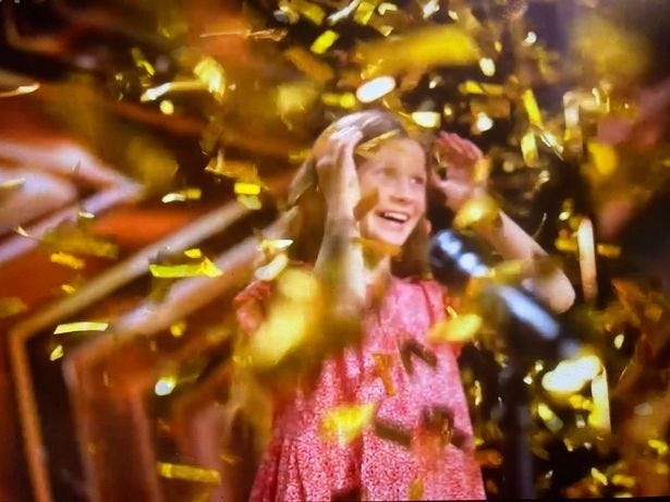 An excited girl in a pink dress in front of a microphone, smiling under a shower of gold confetti An excited girl in a pink dress in front of a microphone, smiling under a shower of gold confetti