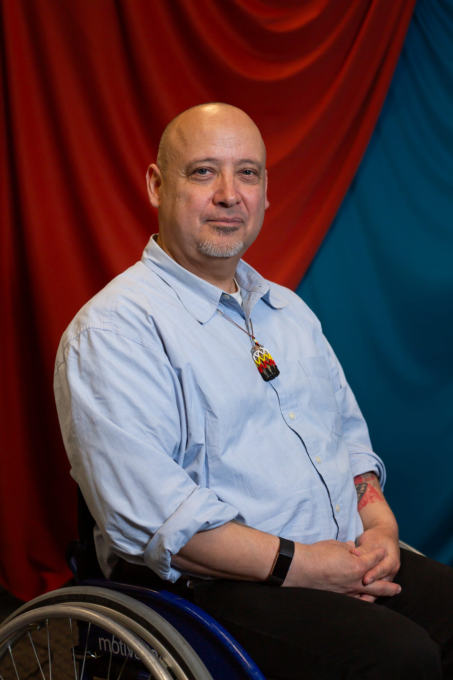 Portrait of a light skinned, early 40’s native man. He is sitting in a wheelchair in front of an orange and teal fabric draped backdrop. He is bald with a grey goatee. He is wearing a light blue collared shirt with the sleeves rolled up. He has a long necklace with a beaded pendant wrapped around his neck. A red and blue tattoo is slightly visible on his left arm.
