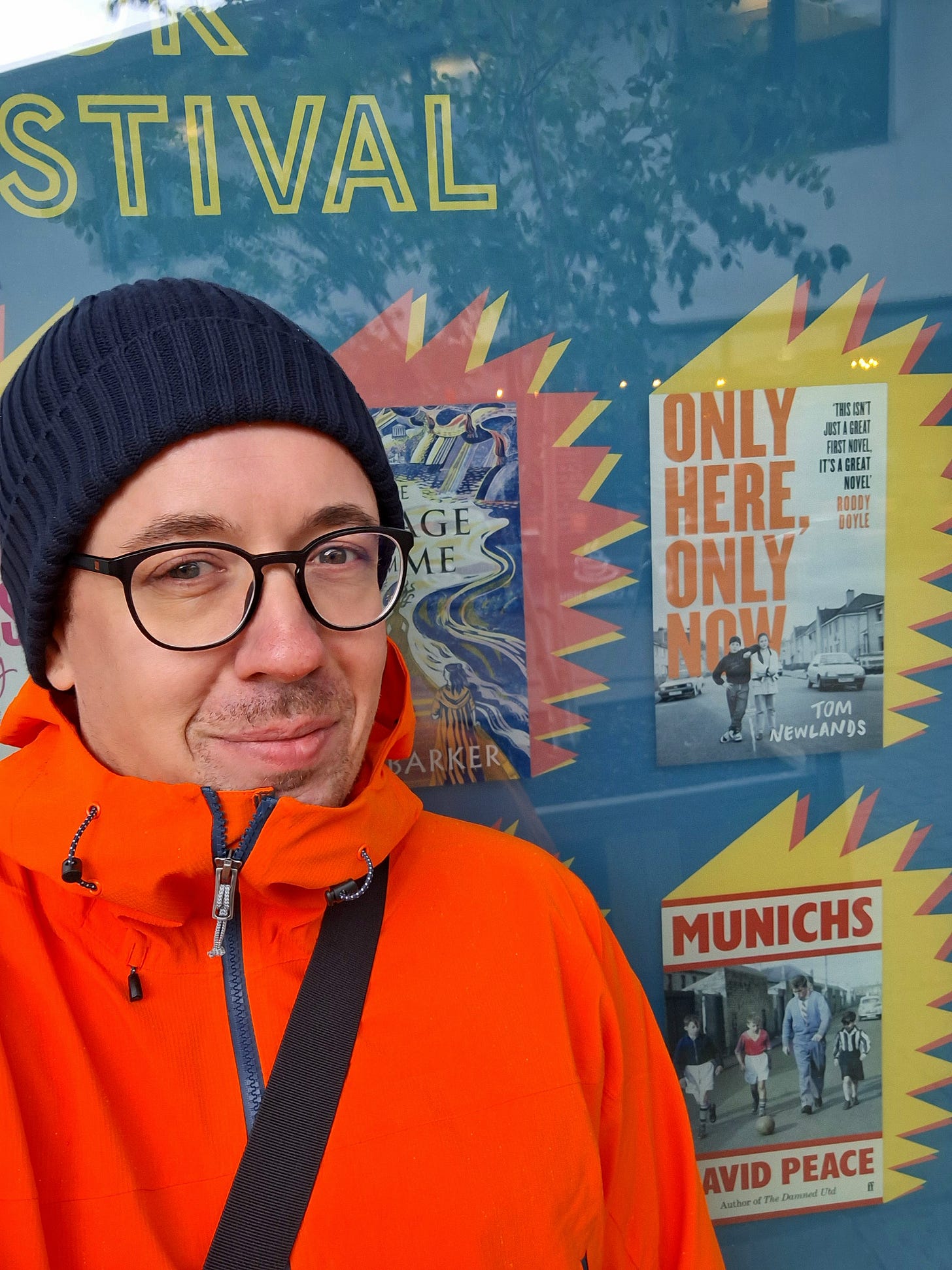 A closeup of a smiling man with spectacles in bright anorak and dark wooly hat, standing in front of a large bright window graphic advertising the book Only Here, Only Now. 