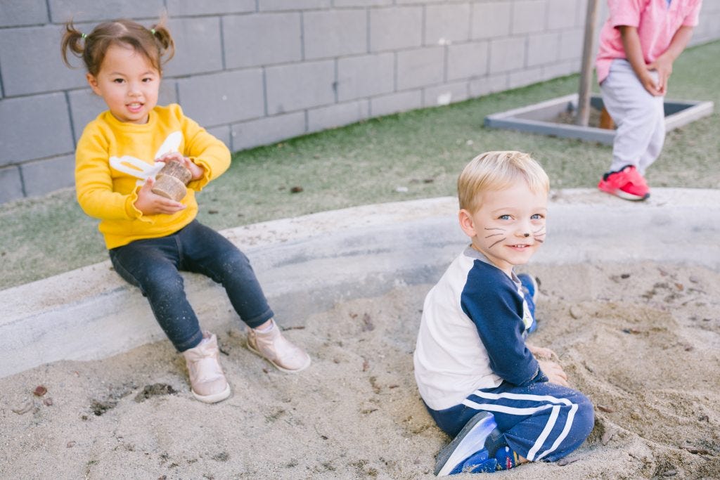Two toddlers sitting in an outdoor sandbox at a Montessori school, one smiling at the camera with face paint