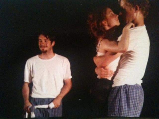 Three young actors stand on stage wearing white t-shirts and checked pyjama bottoms. One stands apart looking out. The other two hug tightly standing up, looking into each other's eyes.  