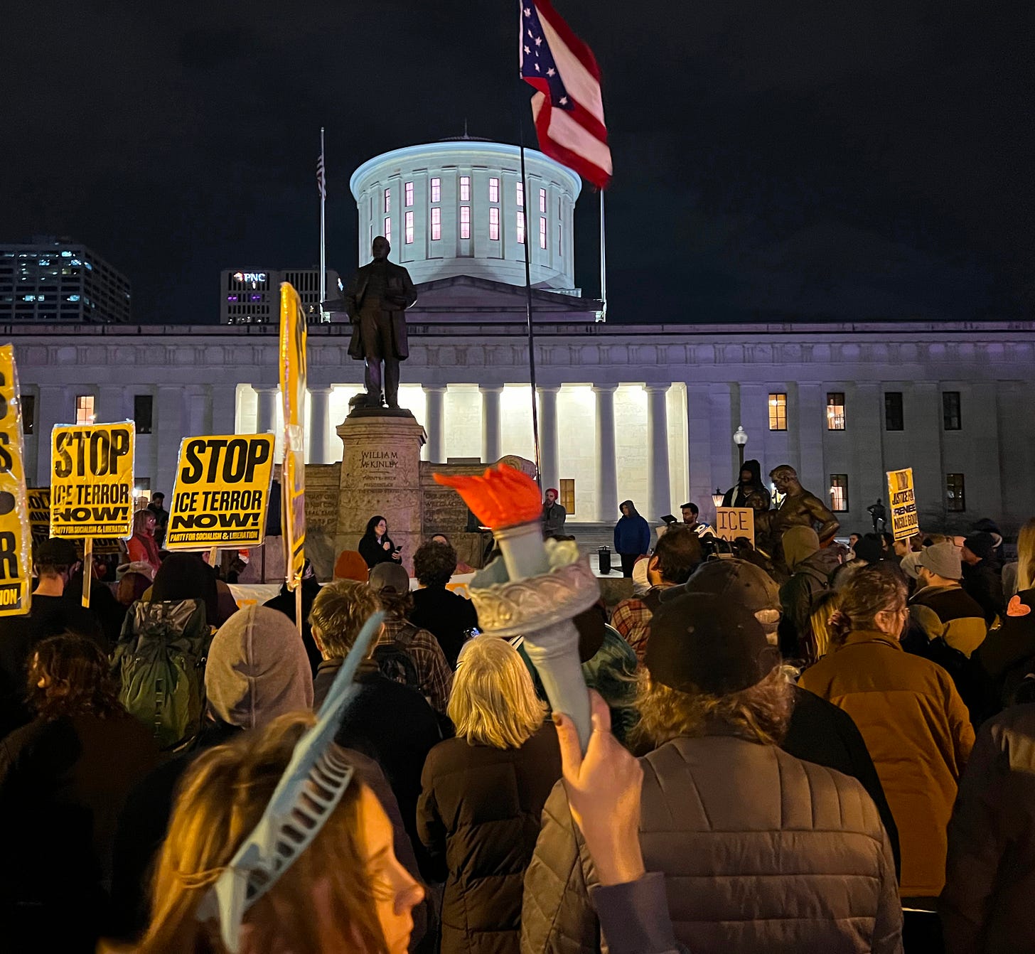 A large group of people bundled up warmly at night in a public plaza. In the background is a large building with classical architecture - the Ohio Statehouse. The protesters are around a statute which is labeled "William McKinley," and a flagpole waving the flag of Ohio. They are holding signs that say things like "STOP ICE TERROR NOW," and one is holding Lady Liberty's torch.