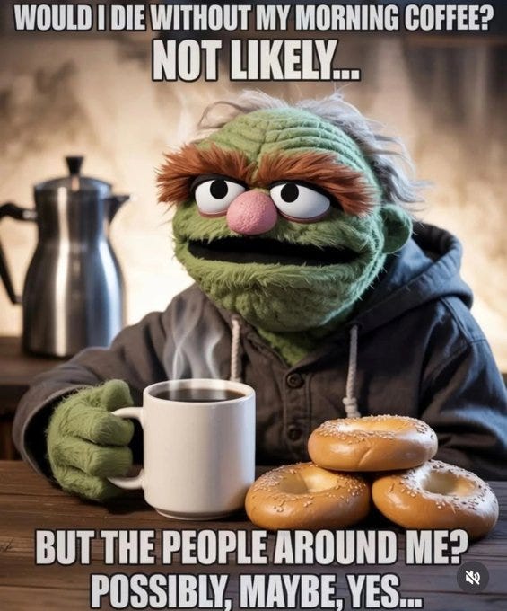 Green furry Muppet character Oscar the Grouch with orange eyebrows and nose sits at a wooden table wearing a gray hooded sweatshirt. He holds a white mug of steaming black coffee in his right hand while looking sideways with a grumpy expression. A silver coffee pot steams on the table to his left. Three golden brown bagels are stacked on a plate next to the mug. Background is warmly lit with blurred neutral tones.