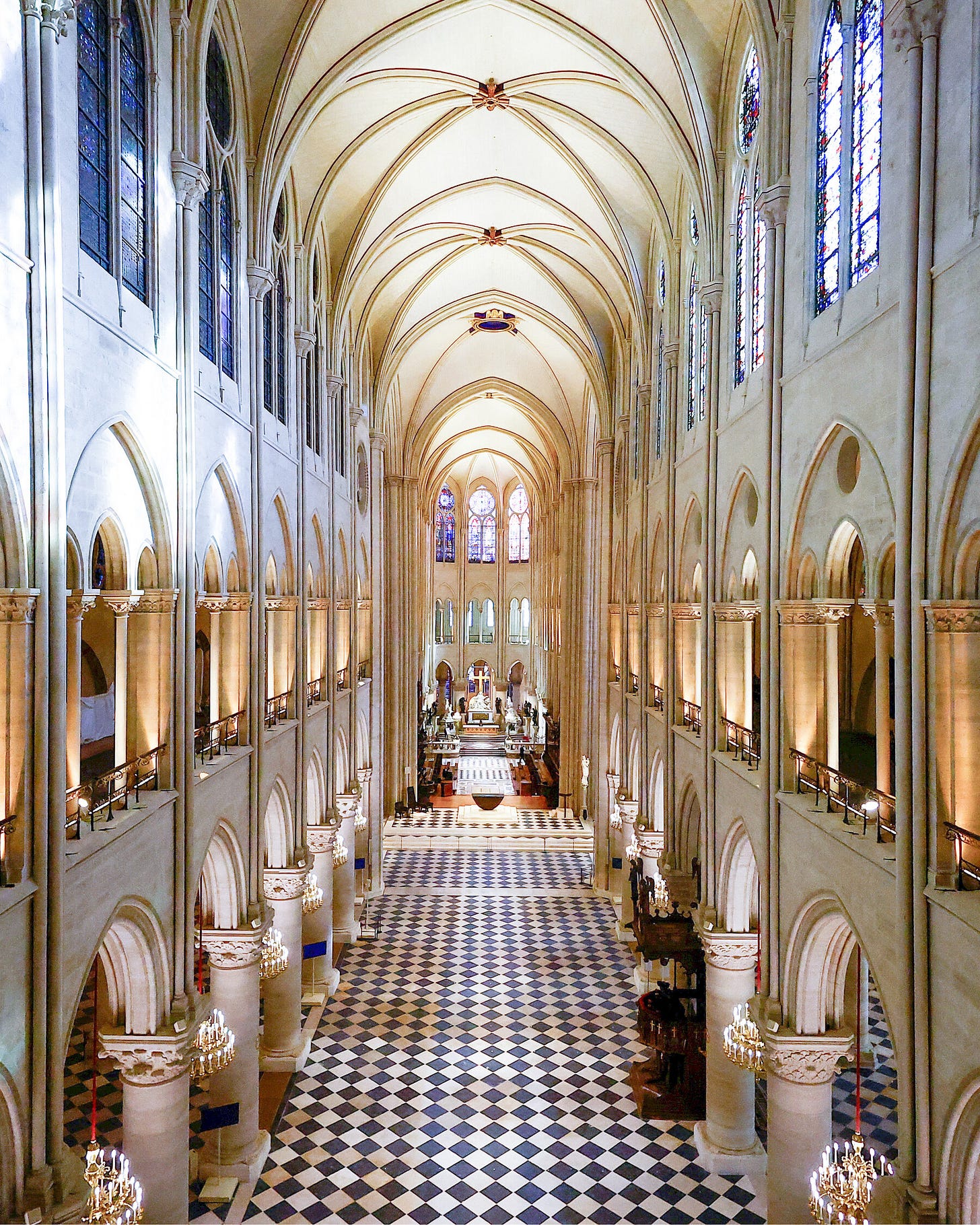 A central view of the nave of Notre-Dame, with a checkerboard floor, arches, columns and stained glass glowing with light.