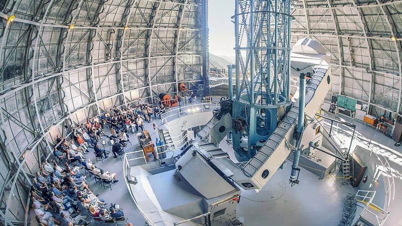 The interior of a massive telescope dome. Only a portion of the spherical walls are visible. The camera’s perspective is hovering above a group of people sitting and gathered around a presentation. The dome’s mechanical walls are partially open, revealing a slice of sunlight sky. The interior of a massive telescope dome. Only a portion of the spherical walls are visible. The camera’s perspective is hovering above a group of people sitting and gathered around a presentation. The dome’s mechanical walls are partially open, revealing a slice of sunlight sky.