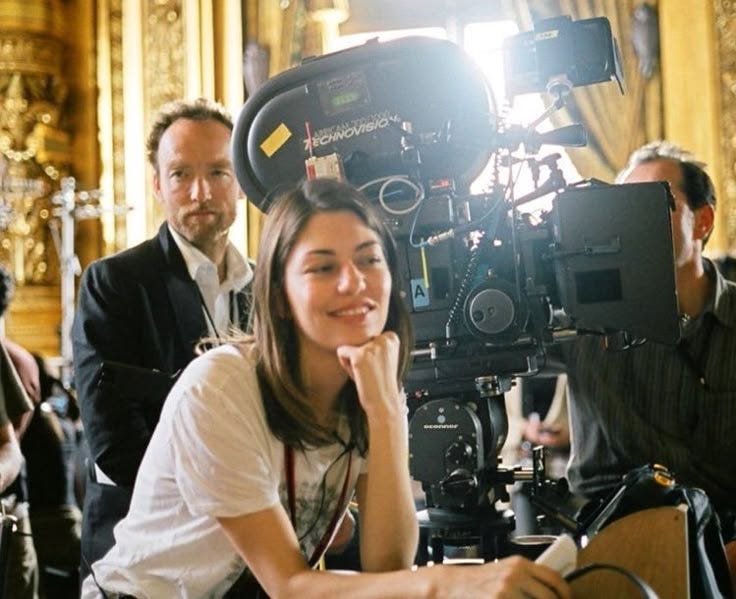 This may contain: a woman sitting in front of a camera on top of a wooden table next to other people