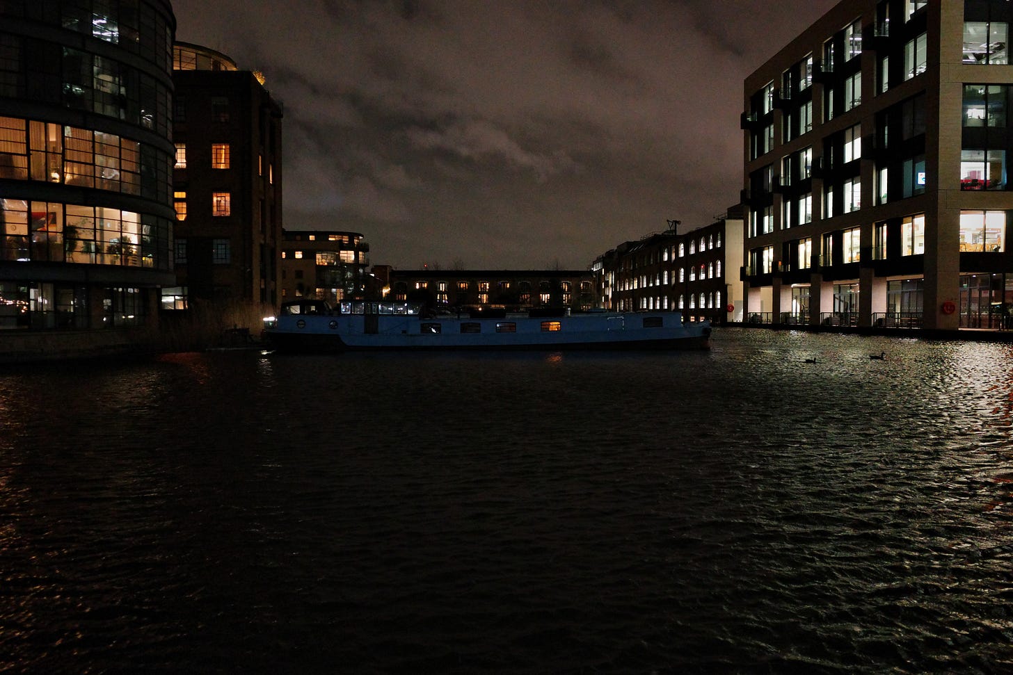 A canal boat rests on the water between a pair of apartment buildings. Reflected light glimmers on the water, making silhouettes of two ducks A canal boat rests on the water between a pair of apartment buildings. Reflected light glimmers on the water, making silhouettes of two ducks