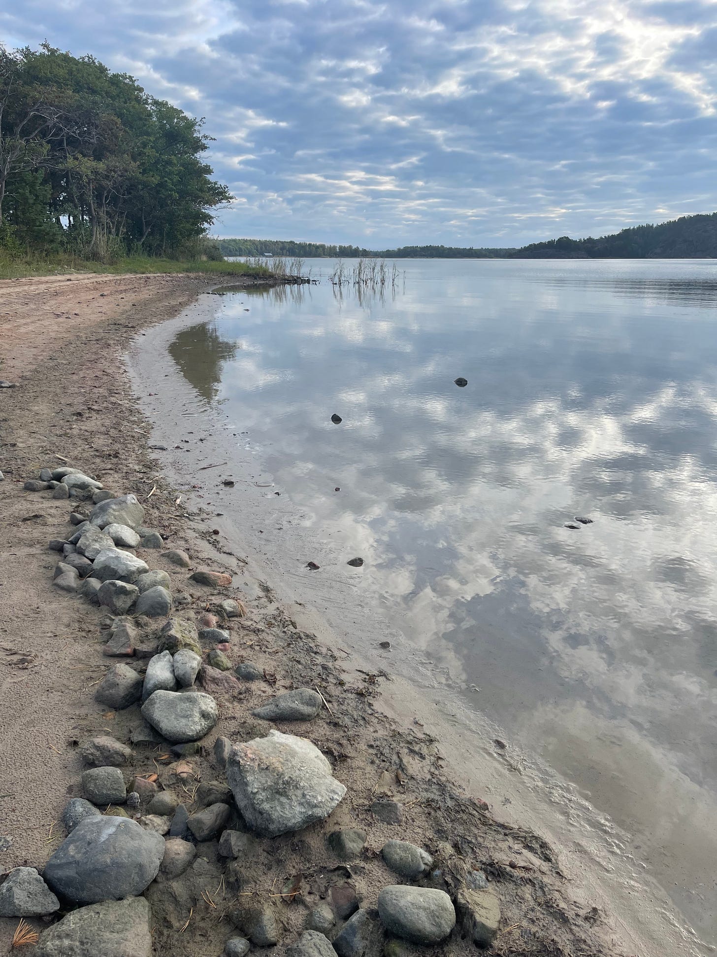 Sky reflected in the water, greys clouds, a strip of sand with some stones