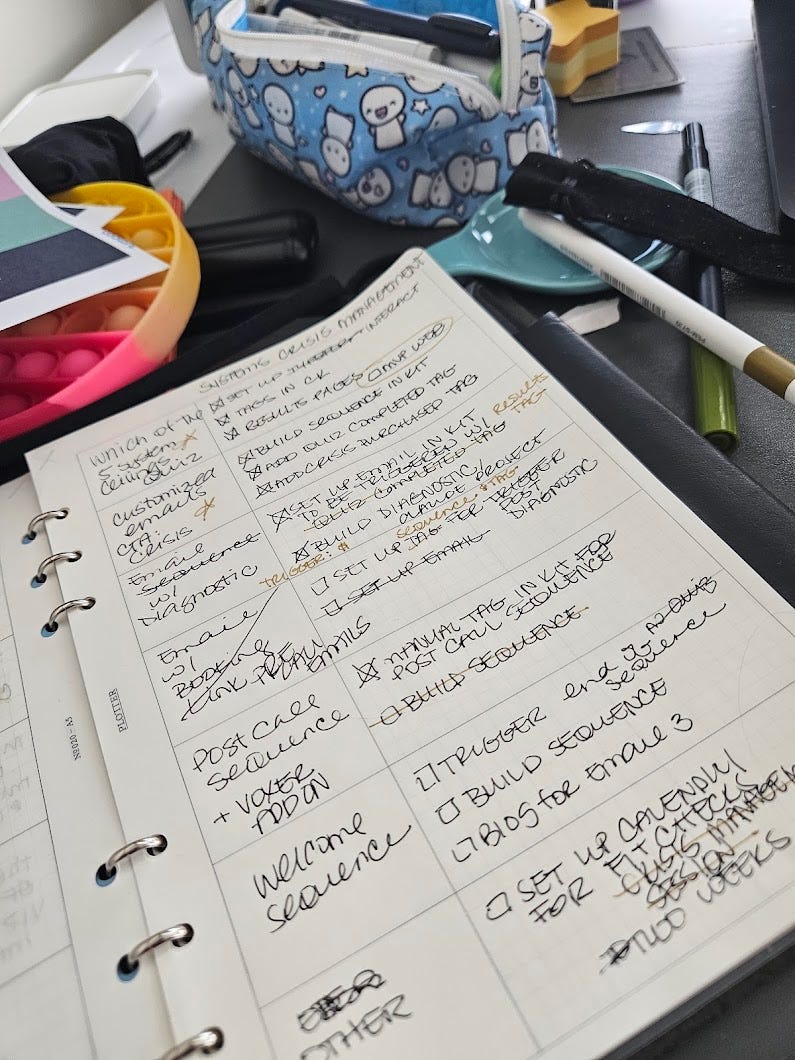 Overhead shot of a paper planner filled with handwritten notes and checkboxes, surrounded by pens, a fidget toy, and a blue Hello Kitty pencil case. The desk is cluttered, creative, and full of energy. Overhead shot of a paper planner filled with handwritten notes and checkboxes, surrounded by pens, a fidget toy, and a blue Hello Kitty pencil case. The desk is cluttered, creative, and full of energy.