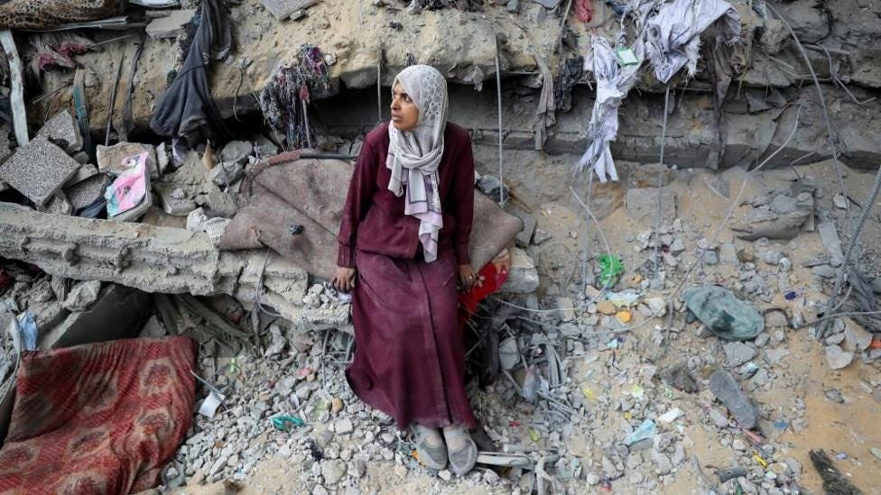 A woman wearing a purple dress and grey headscarf sits on a blanket amidst a pile of rubble.