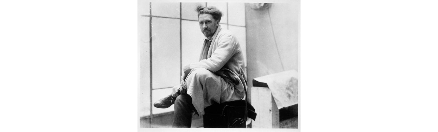 Ezra Pound sitting at a window with one leg crossed over the other, in a white coat, his wiry hair uncombed