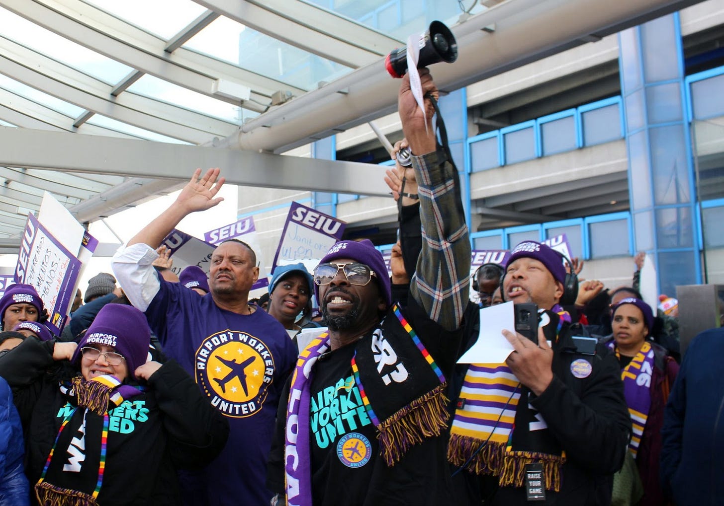 a crowd of people of color wearing purple hats and holding up purple SEIU picket signs, one black man in the center wearing sunglasses holds up a small black megaphone a crowd of people of color wearing purple hats and holding up purple SEIU picket signs, one black man in the center wearing sunglasses holds up a small black megaphone