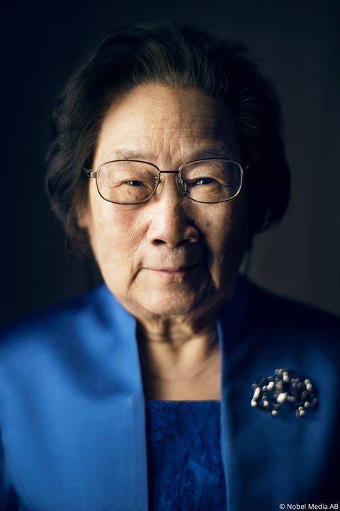 A portrait of Tu Youyou in front of a dark grey background. The light is on the left side of her face leaving the right side slightly in shadow. She is looking directly into the camera with a small smile.