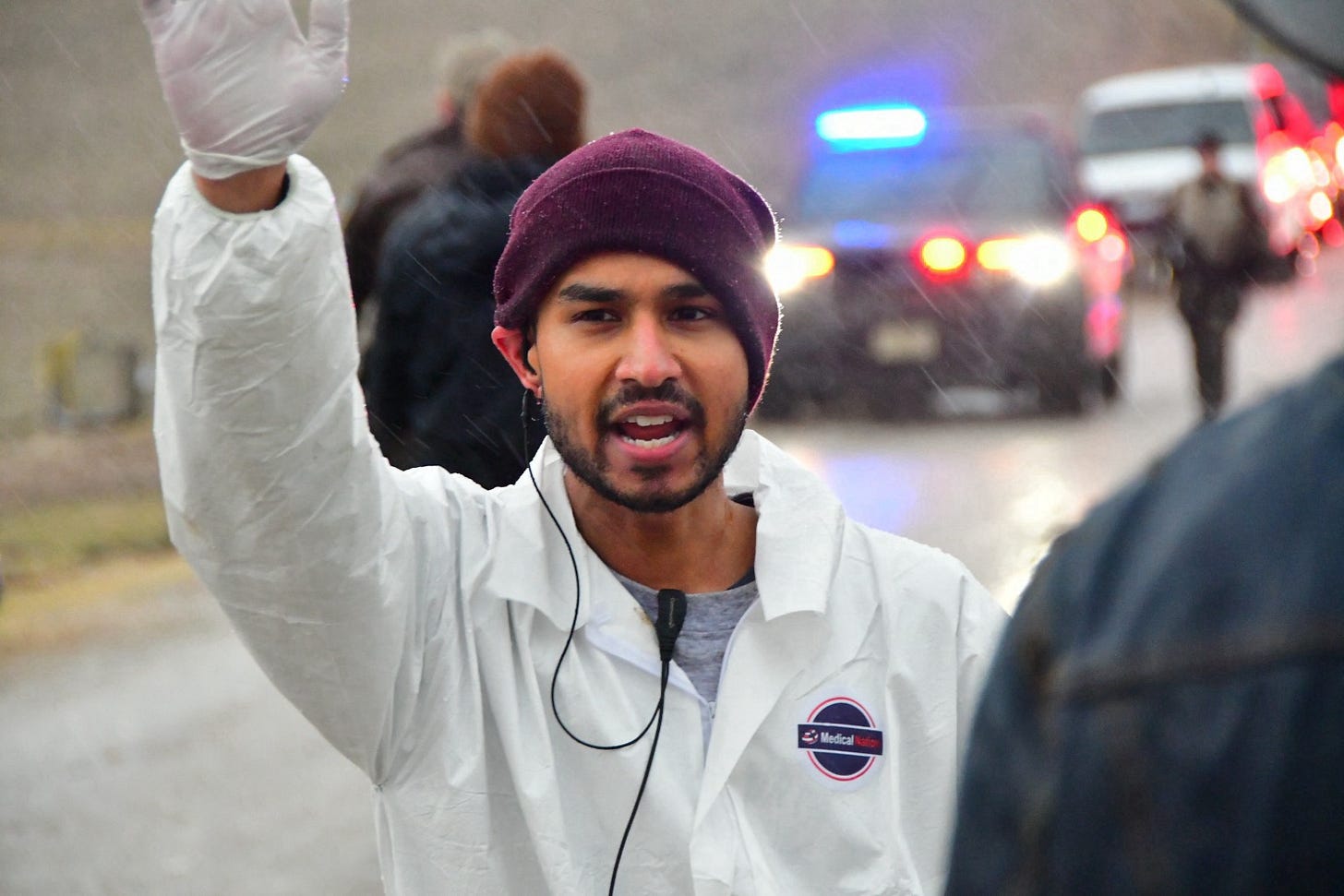 Close-up of an activist in a white coverall and maroon beanie raising a gloved fist in the rain. Police vehicles with flashing red and blue lights are visible in the background. The activist wears a 'Medical' badge on their coverall.