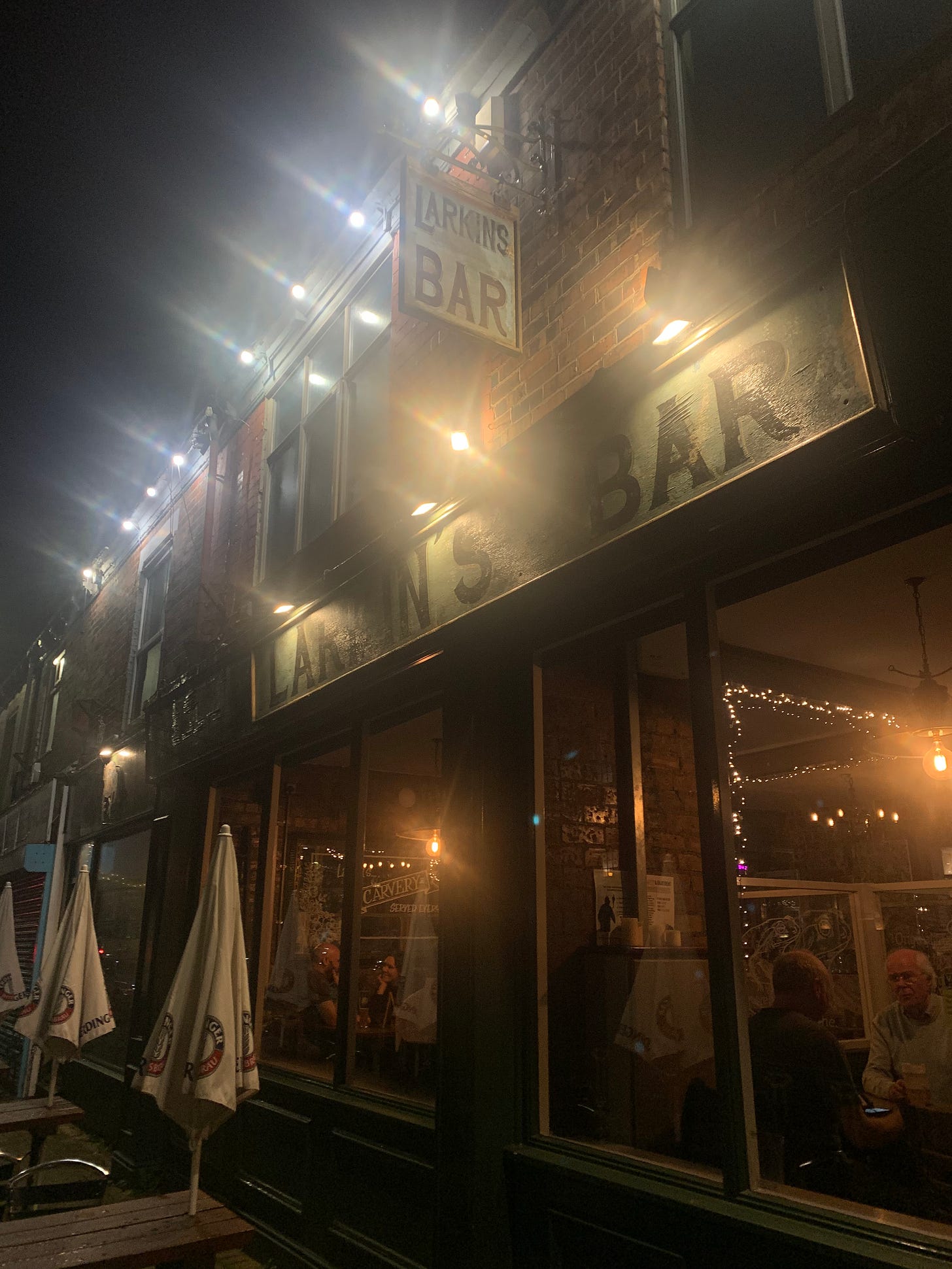 A photo of outside Larkin's Bar in Hull. It's night time and there's a sign that says Larkins Bar, the photo looks into the bar where you can see people sitting and chatting through the windows. A photo of outside Larkin's Bar in Hull. It's night time and there's a sign that says Larkins Bar, the photo looks into the bar where you can see people sitting and chatting through the windows.