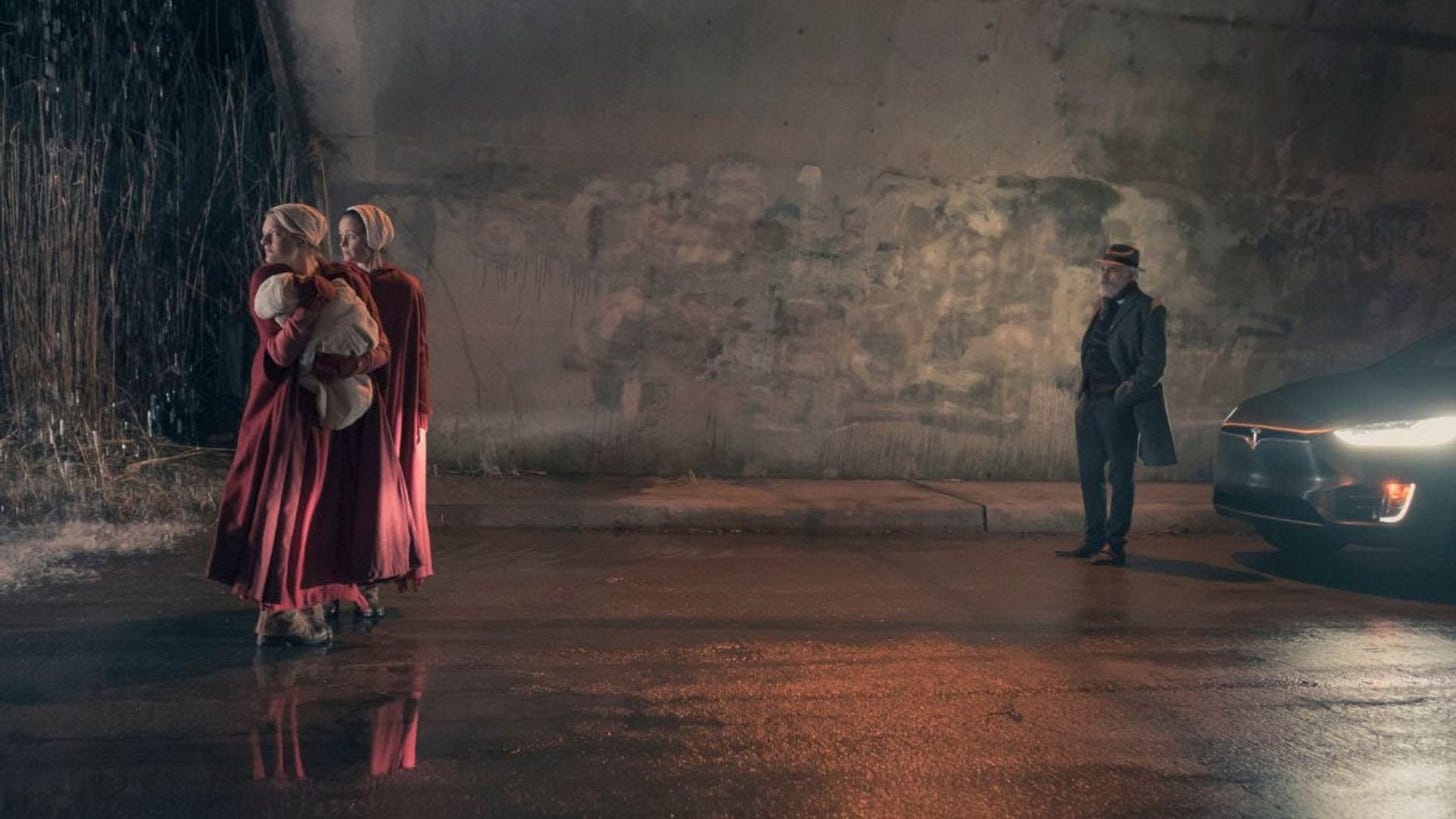 Screenshot from Handmaid's Tale TV show with Commander Lawrence, a middle-aged white man in a suit, and two women dressed as handmaids, one holding a baby. They are in a tunnel with a Tesla with its lights on