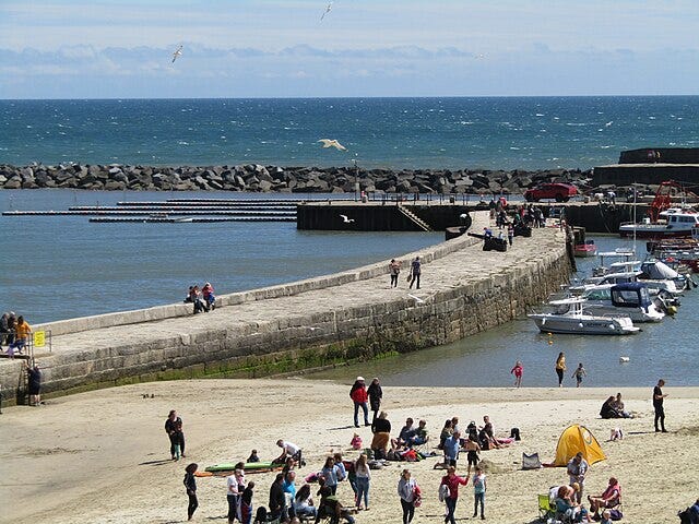 Photo by Colin Smith of tourists lounging on the beach with the Lyme Regis Breakwater in the background. A curving causeway built of stone rises above placid water leading to a marina and eventually the breakwater. In the background beyond, the water is white capped and choppy.