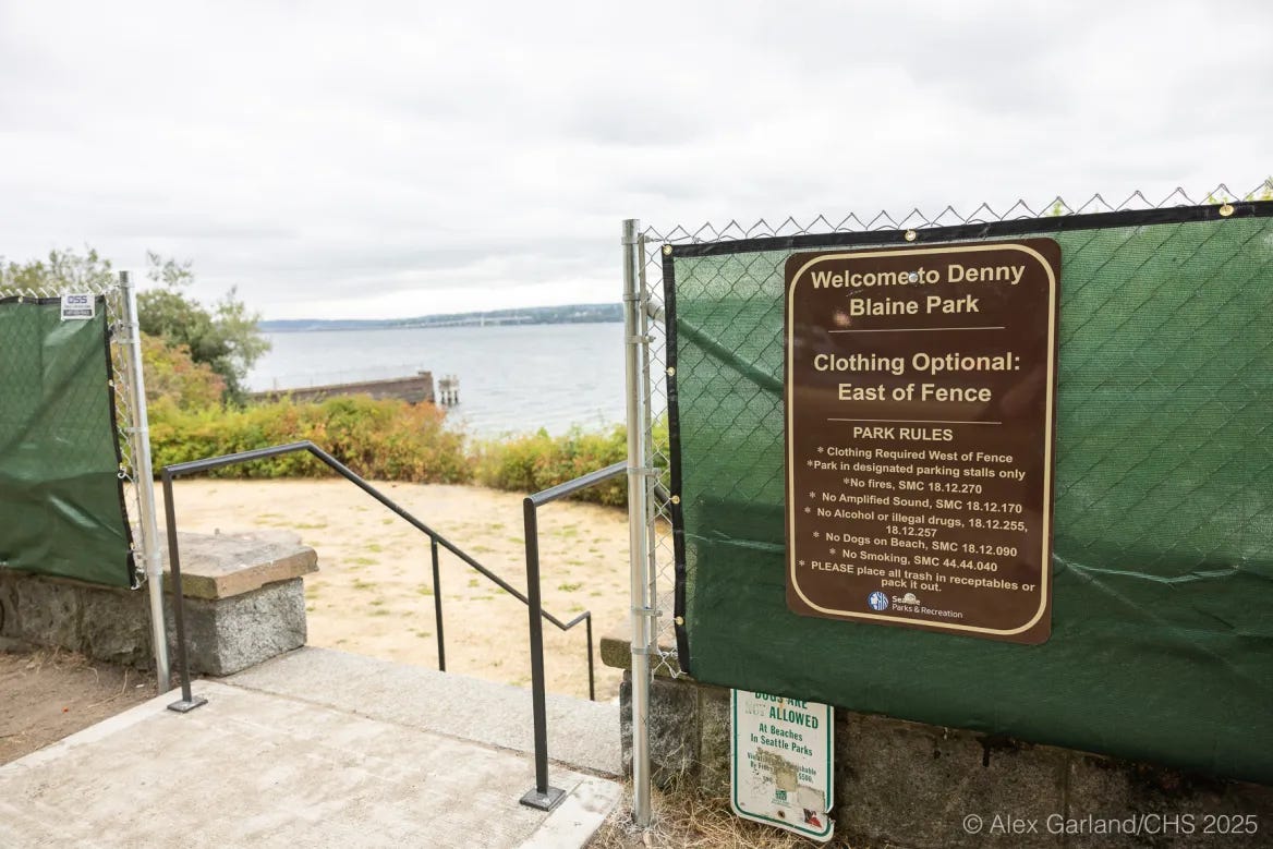 A green chain-link fence with tarps and a brown Seattle Parks sign reading “Clothing Optional: East of Fence” stands at the entrance to Denny Blaine Park overlooking Lake Washington. A green chain-link fence with tarps and a brown Seattle Parks sign reading “Clothing Optional: East of Fence” stands at the entrance to Denny Blaine Park overlooking Lake Washington.