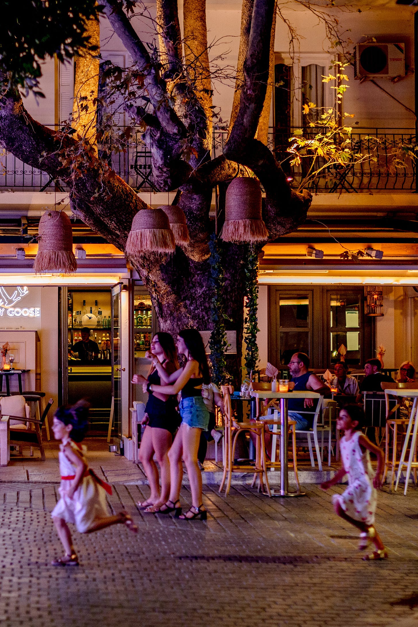 Adults and children make their way down a street and past a restaurant with outdoor tables at nighttime.