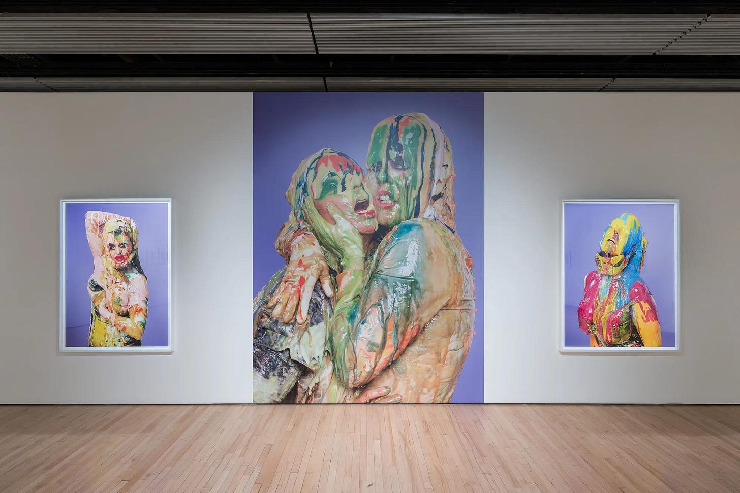 Three large artworks installed on a white wall, each feature a figure covered in multicolours paint on a purple background