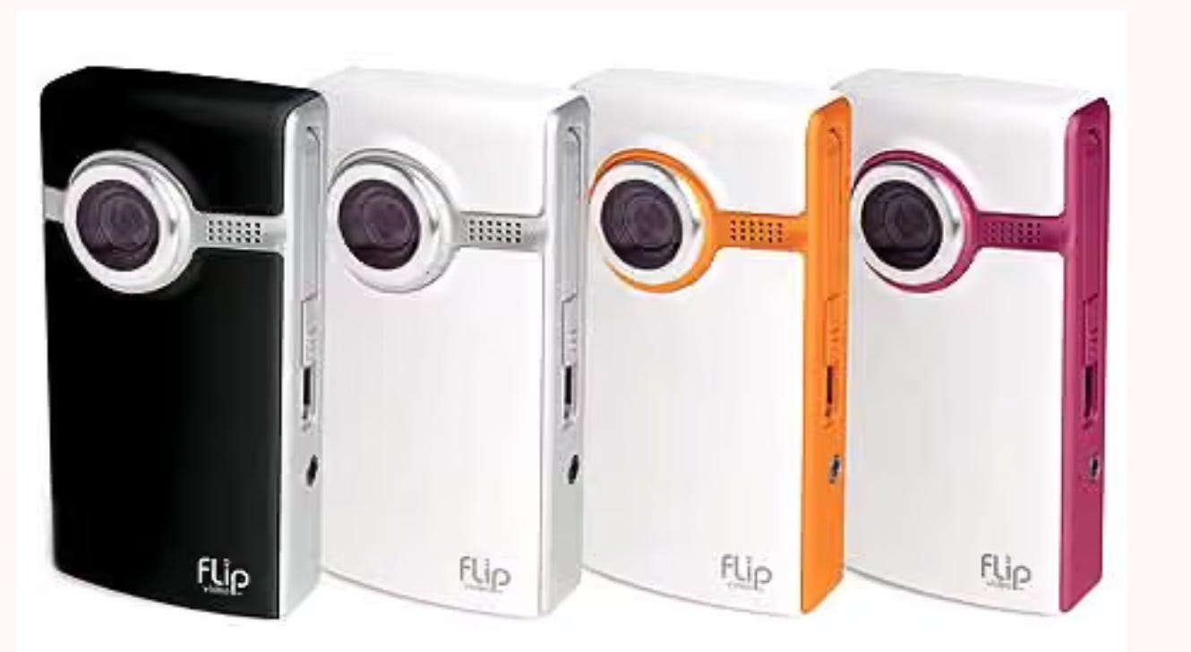 Four Flip Video cameras standing upright in a row—black, white, orange, and pink—showing the simple, early-era device I used to film my first unpolished blogging videos.