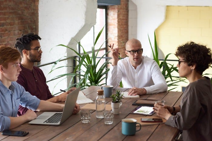 Several people sitting at a coffee shop. One guy in the center is gesturing with his hand, perhaps as a facilitator. The two people on the left are taking notes through journals or computers, and the woman on the right may be explaining something as a participant.