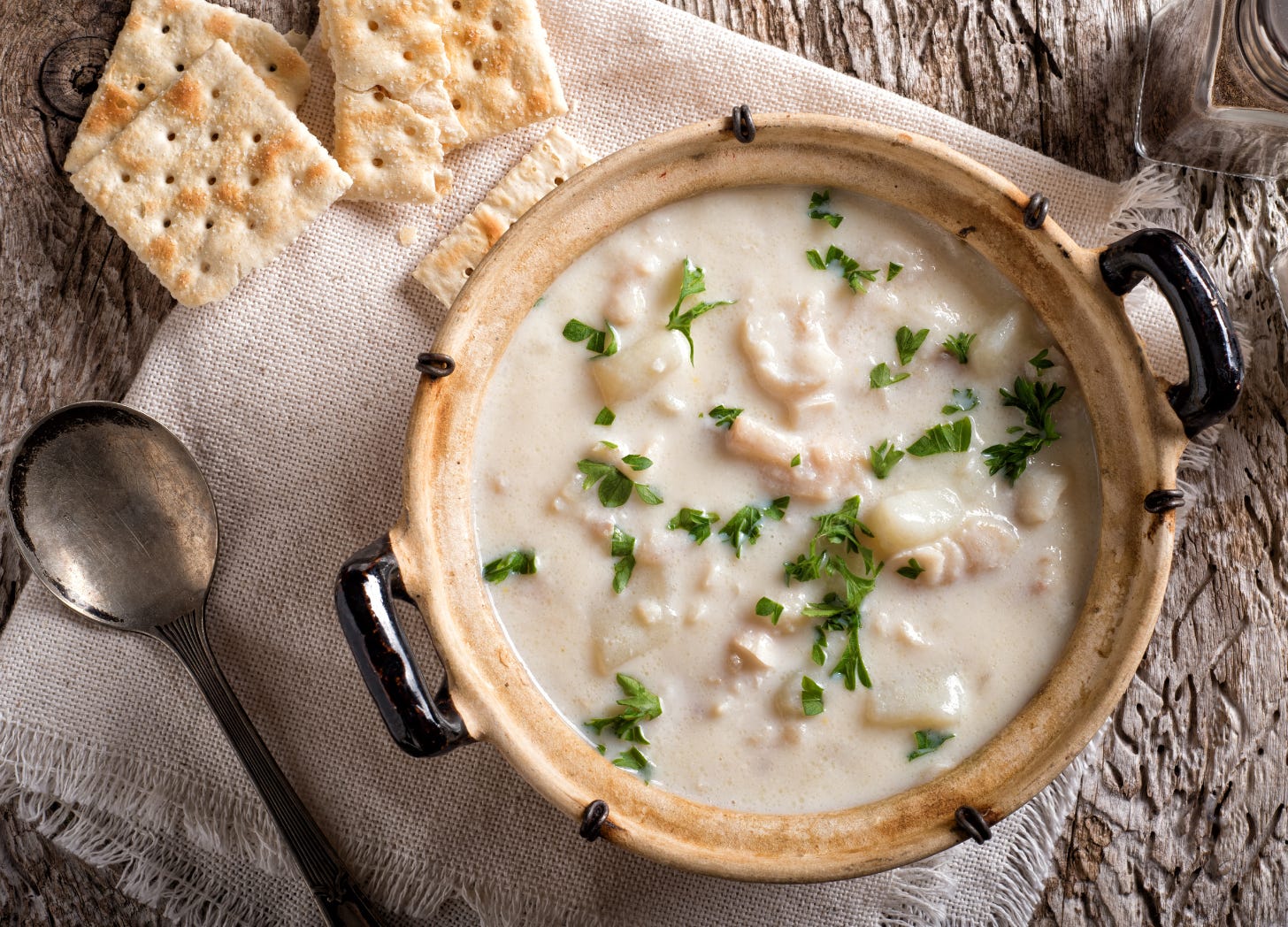 a bowl of seafood chowder sits on a table with a rustic cloth napkin, a well worn soup spoon and four saltine crackers, one is cracked and it looks like someone is about to start enjoying the bowl of soup.