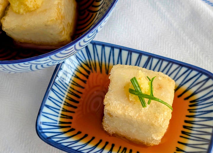 Agedashi Tofu in Light Soy-Based Sauce: Crispy, golden-brown cubes of agedashi tofu sit in a shallow pool of light, savory soy-based sauce. The dish is garnished with finely sliced scallions and freshly grated ginger, adding a pop of color and freshness to the delicate, crispy tofu.