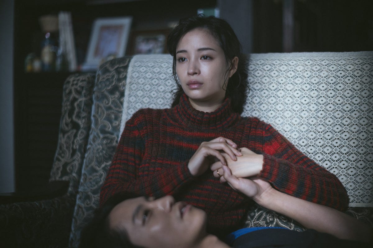 A imagem mostra Sakiko vestindo uma blusa de gola alta vermelha e preta listrada e com uma expressão melancólica, sentada em um sofá com estampado claro. Ela está segurando as mãos de seu companheiro, deitado com a cabeça em seu colo.