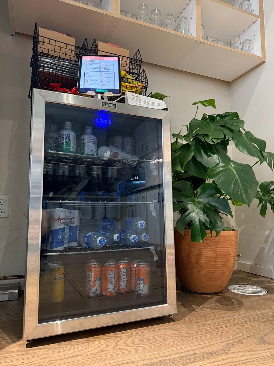 The physical setup of Project Vend: a small refrigerator, some stackable baskets on top, and an iPad for self-checkout.
