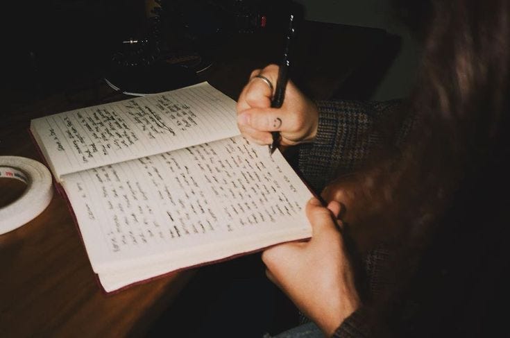 This may contain: a person sitting at a table with a book and pen in their hand, writing on it