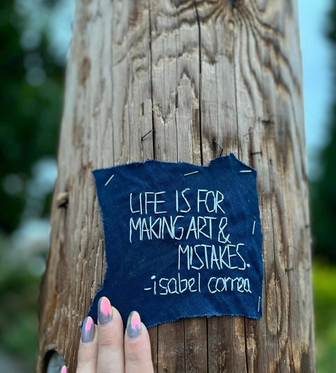 The image features a piece of embroidered fabric attached to a wooden post, with the text "LIFE IS FOR MAKING ART & MISTAKES" written in white thread, attributed to Isabelle Correa. This artwork, embroidered by Shana Lee Hampton, is a reflection of the theme from Isabelle Correa's poem September as Strip Tease. The piece showcases a thoughtful and creative exploration of the relationship between imperfection and art. A hand with multi-colored nails gently touches the fabric, emphasizing the personal and tactile connection to the message.