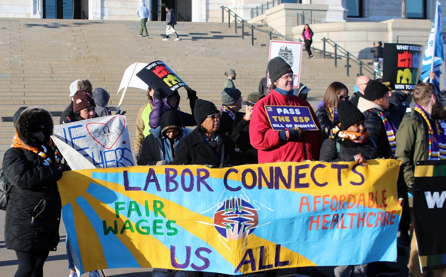 people standing with colorful signs and wearing winter attire hold a banner depicting a blue sky and yellow sun background behind text that reads "labor connects us all" "Fair wages" and. "affordable healthcare" with 4 hands reaching fingertips people standing with colorful signs and wearing winter attire hold a banner depicting a blue sky and yellow sun background behind text that reads "labor connects us all" "Fair wages" and. "affordable healthcare" with 4 hands reaching fingertips